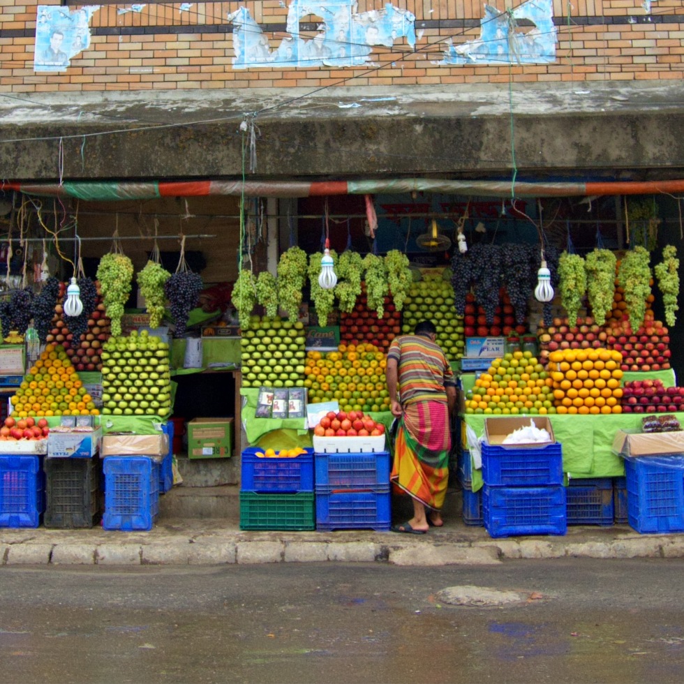 Three stalls selling a wide variety of fruit. Each type of fruit is stacked in to beautiful neat pyramids. Bunches of juicy grapes hang above each stall.